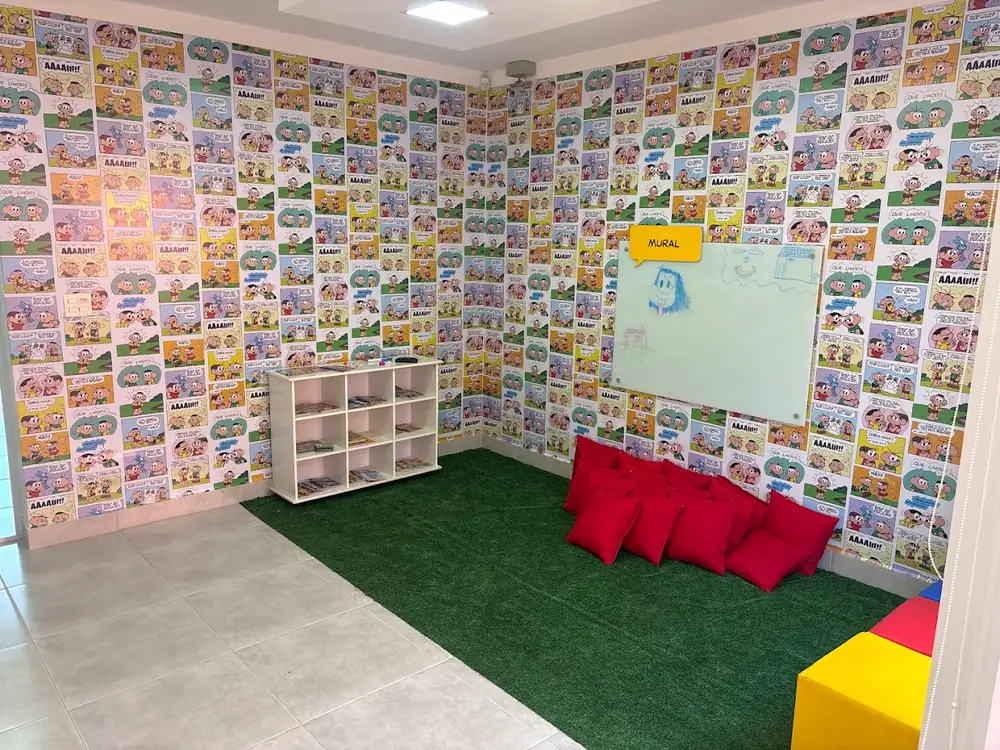 Sala do Saber com tapete verde, almofadas, estante de livros e mural